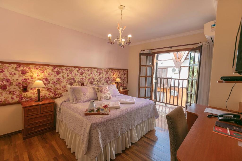 Uma cama bem arrumada com roupa de cama floral, uma escrivaninha de madeira e portas de vidro abertas que dão para uma varanda em um quarto bem iluminado, na Pousada das Hortênsias, para representar pousadas em Campos do Jordão.