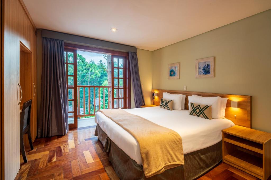Um quarto na Pousada Campos dos Holandeses com uma cama grande, mesas de cabeceira, piso de madeira, escrivaninha e uma varanda com vista para a vegetação, para representar pousadas em Campos do Jordão.