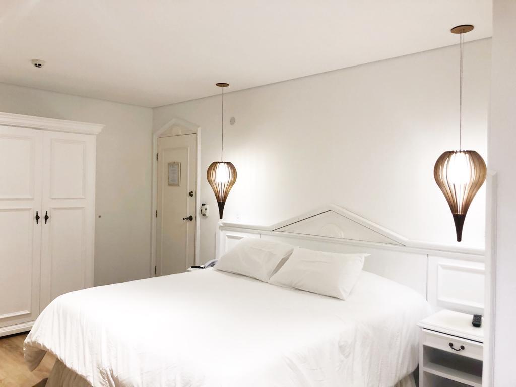 Um quarto branco com uma cama de casal, dois pendentes, um guarda-roupa e um criado-mudo contra uma parede branca, na Pousada Dom Alfredo José, para representar pousadas em Campos do Jordão.