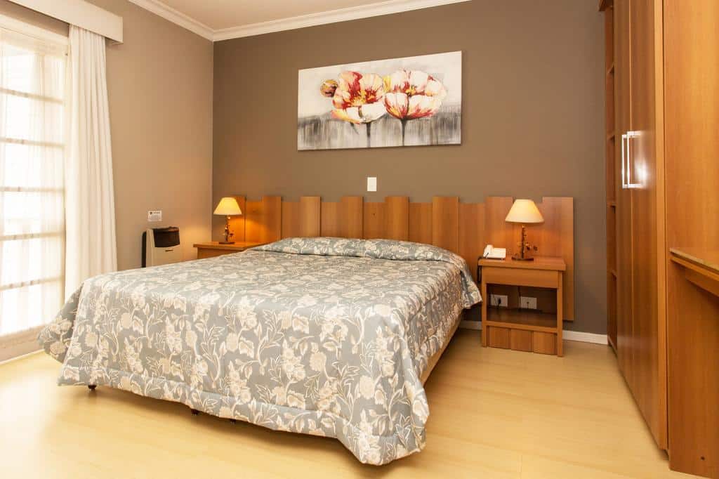 Um quarto na Pousada Nacional Inn com uma cama de casal, duas mesas de cabeceira com luminárias, móveis de madeira e uma pintura floral acima da cama, para representar pousadas em Campos do Jordão.