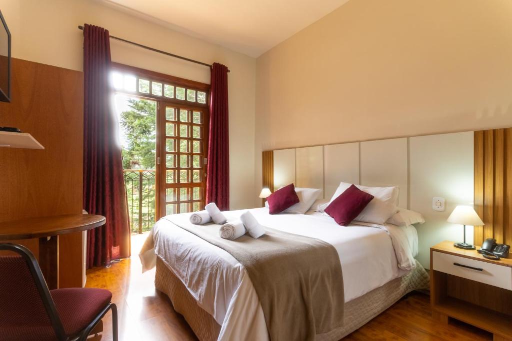 Um quarto na Pousada La Toscana com uma cama de casal, duas mesinhas de cabeceira, luminárias, uma mesa, uma cadeira e portas de vidro que se abrem para uma vista do jardim, para representar pousadas em Campos do Jordão.