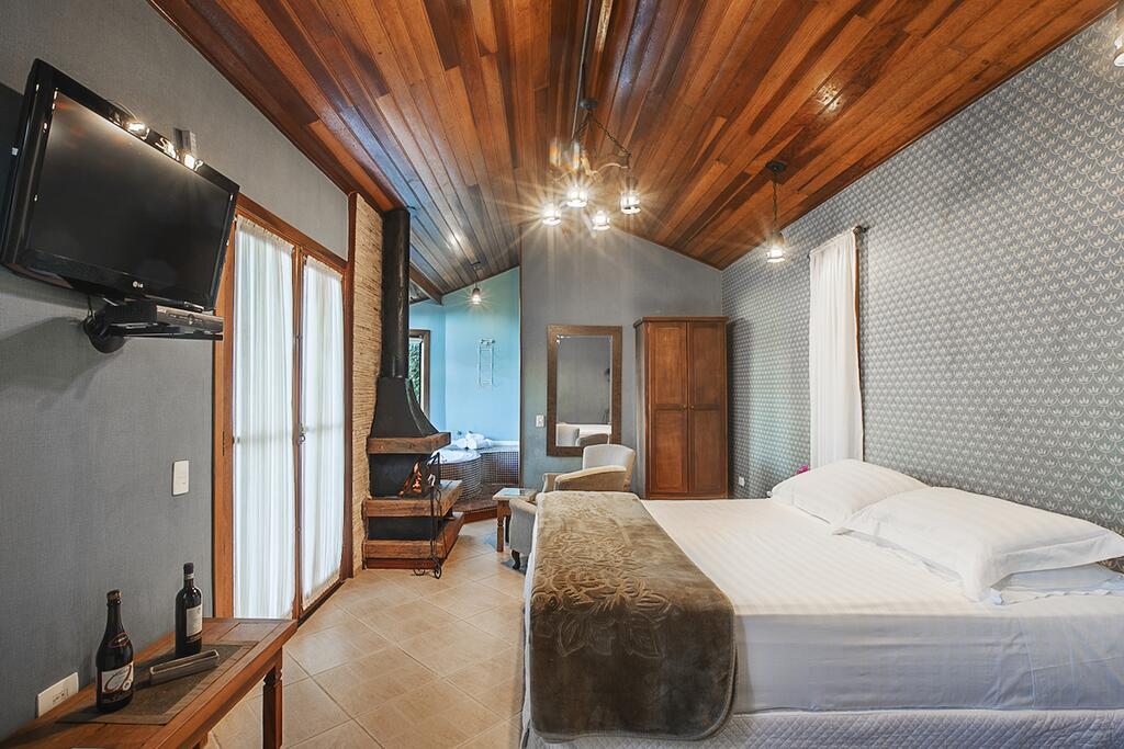 Um quarto com uma cama de casal, TV na parede, teto de madeira, guarda-roupa e uma pequena área de estar com lareira, no Recanto Vila das Flores, para representar pousadas em Campos do Jordão.