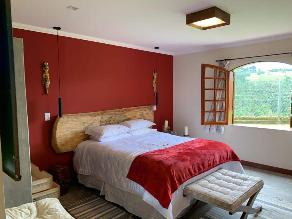 Uma cama bem arrumada com roupa de cama vermelha e branca fica encostada em uma parede com detalhes vermelhos em um quarto com uma janela aberta, na Villa Capuzzo, para representar pousadas em Campos do Jordão.