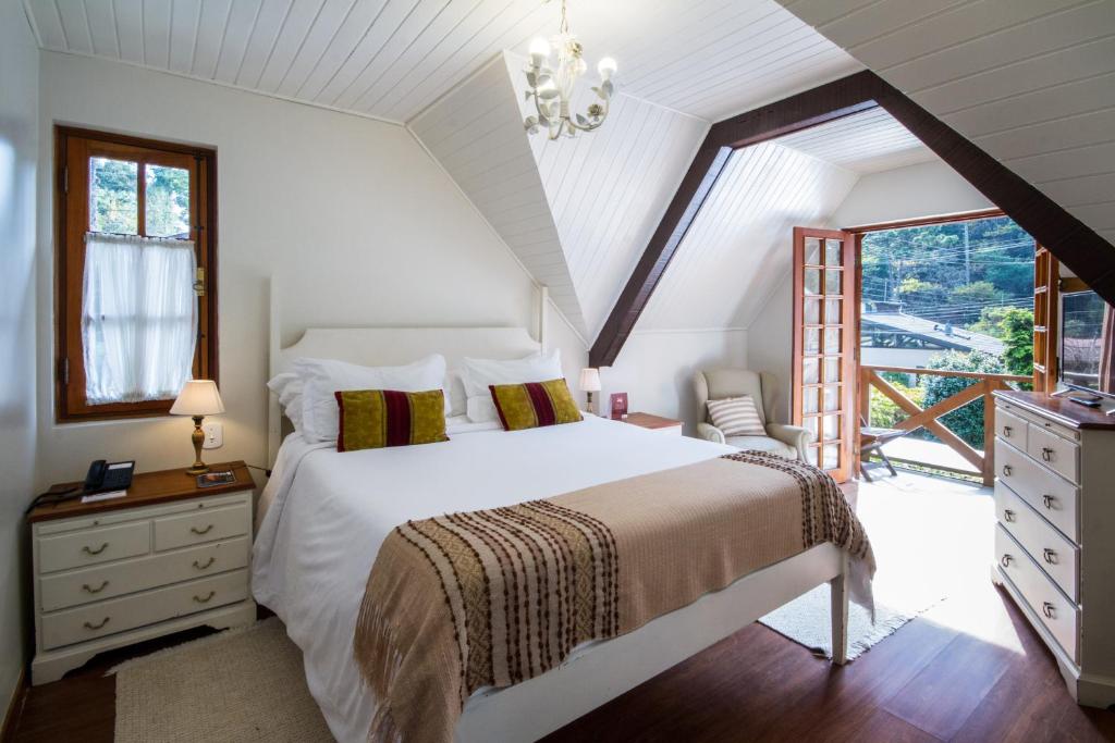 Um quarto na Pousada da Pedra com uma cama grande, duas mesinhas de cabeceira, poltrona, cômoda e portas abertas para uma varanda com vista, para representar pousadas em Campos do Jordão.