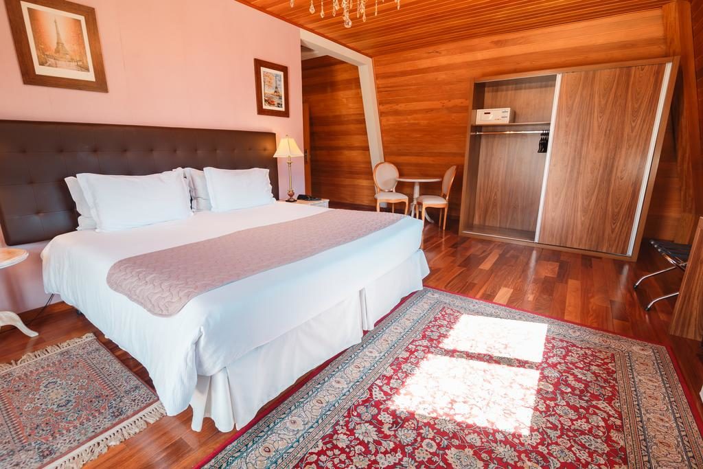 Um quarto na Pousada Luis XV com uma cama de casal, piso de madeira, dois tapetes, um guarda-roupa e uma pequena mesa com duas cadeiras, para representar pousadas em Campos do Jordão.