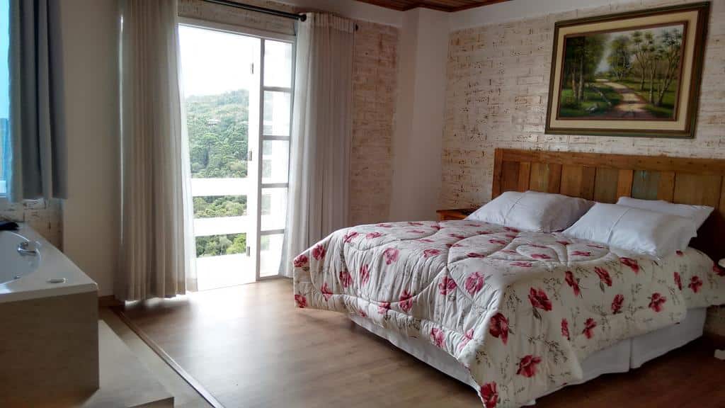 Um quarto na Pousada Miraggio com colcha floral, cabeceira de madeira, arte na parede, janela grande e uma banheira de hidromassagem perto da porta, para representar pousadas em Campos do Jordão.