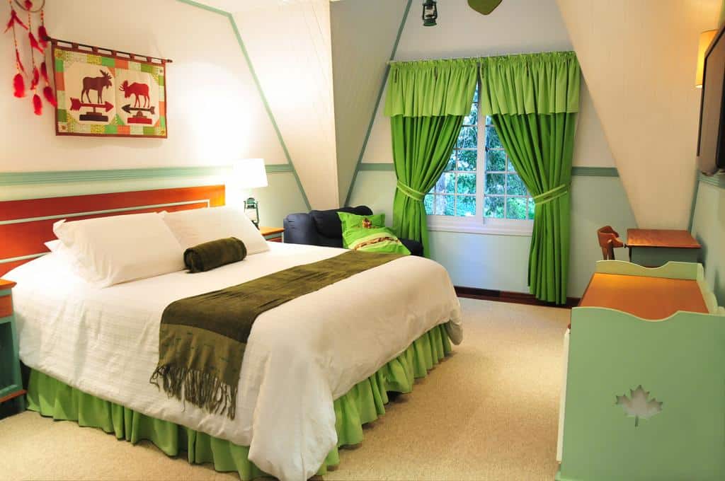 Quarto no Canada Lodge Campos do Jordão com uma cama grande, cortinas verdes, roupa de cama verde e branca, uma janela e uma pequena escrivaninha com uma cadeira.