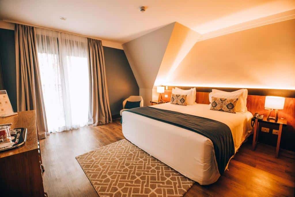 Um quarto no Villa Amistà Campos do Jordão com uma cama grande, duas mesas de cabeceira com luminárias, um tapete estampado e uma janela com cortinas.