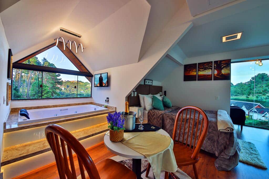 Um quarto aconchegante na Pousada Caminho das Pedras com uma cama de casal, mesa redonda, cadeiras, uma banheira grande e uma janela com vista para árvores e casas, para rperesentar pousadas em Campos do Jordão.