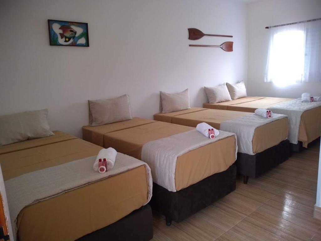 Quatro camas de solteiro com roupa de cama bege estão alinhadas em uma fileira em um quarto bem iluminado com dois remos e uma pintura de peixe na parede, na Pousada Parapuã (Massaguaçu), para representar pousadas em Caraguatatuba.
