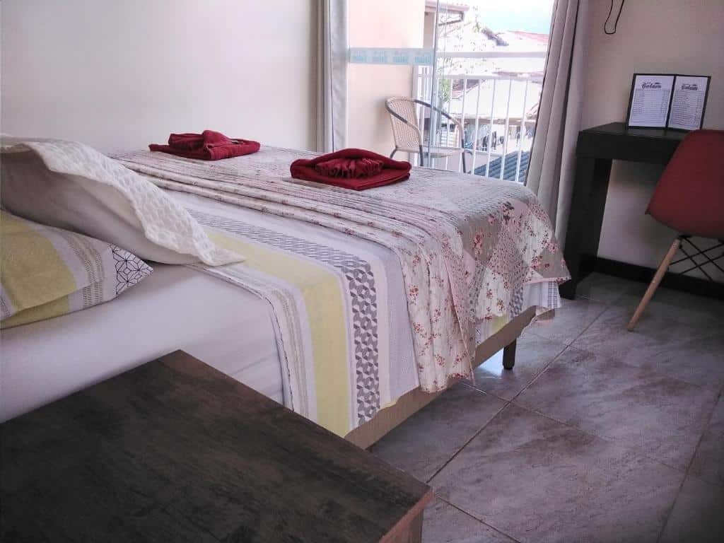 Uma cama bem arrumada com toalhas vermelhas dobradas, uma pequena escrivaninha, uma cadeira e uma varanda do lado de fora de uma porta de vidro, na Pousada Gotam (Aruan/Indaiá), para representar pousadas em Caraguatatuba.