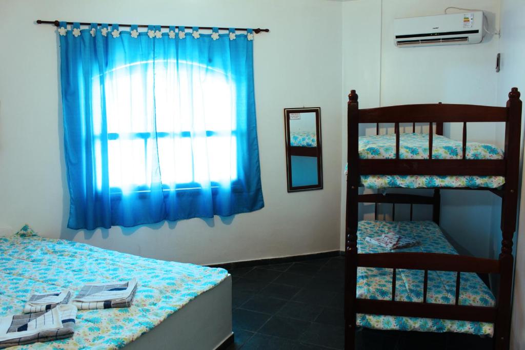 Quarto com uma cama de casal, beliche, roupa de cama com estampa azul, cortinas azuis, espelho de parede e ar-condicionado, no Farol das Gaivotas Pousada e Residence (Martim de Sá), para representar pousadas em Caraguatatuba.