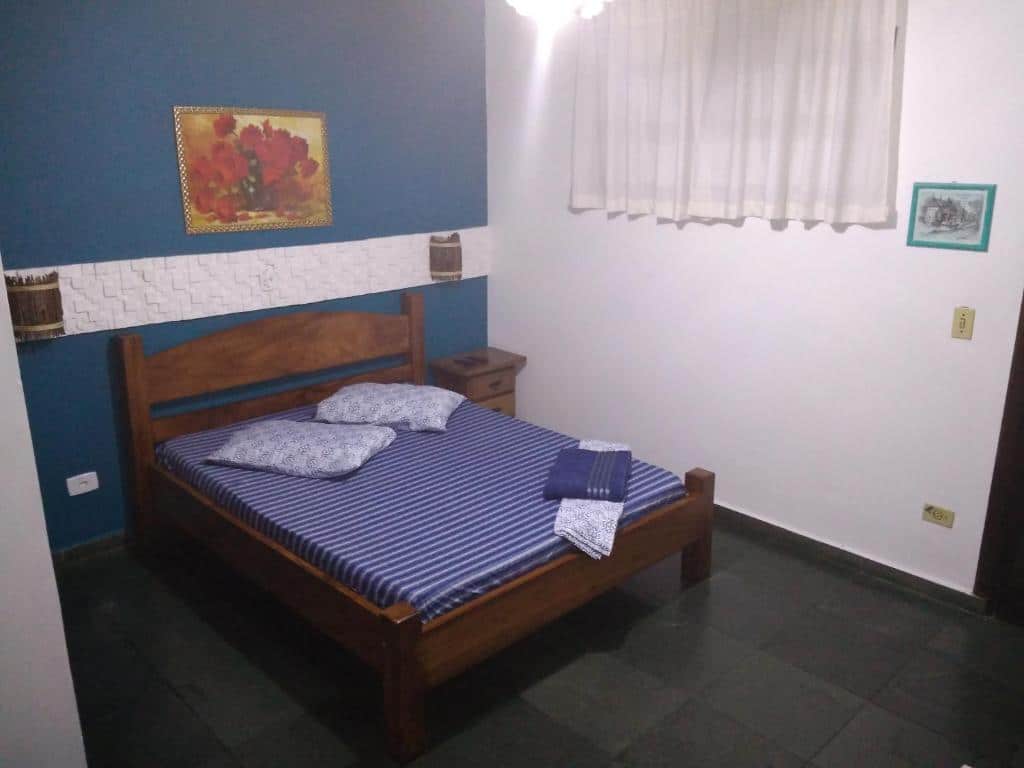 Quarto com cama de casal, luminárias na parede atrás da cabeceira, quadro de flores e uma janela com cortina, na Pousada Costa Verde (Tabatinga), para representar pousadas em Caraguatatuba.