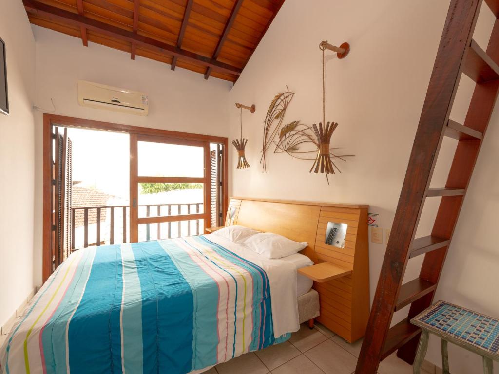 Quarto pequeno com uma cama de casal, roupa de cama listrada, teto de madeira, luminárias de parede, escada e portas de varanda, na Pousada Pé Na Areia (Capricórnio I), para representar pousadas em Caraguatatuba.