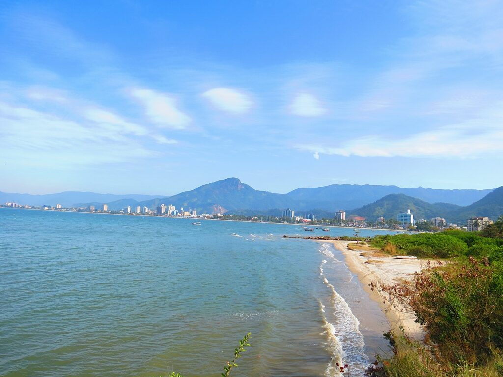 Costa litorânea com ondas suaves, vegetação verde, prédios urbanos distantes e montanhas sob um céu azul, na Orla de Caraguá vista do alto do Camaroeiro.