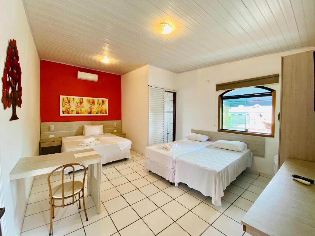 Quarto na Pousada Porto do Rio Praia (Porto Novo) com duas camas, uma mesa com uma cadeira, uma janela, iluminação clara e uma parede vermelha com obras de arte, para representar pousadas em Caraguatatuba.