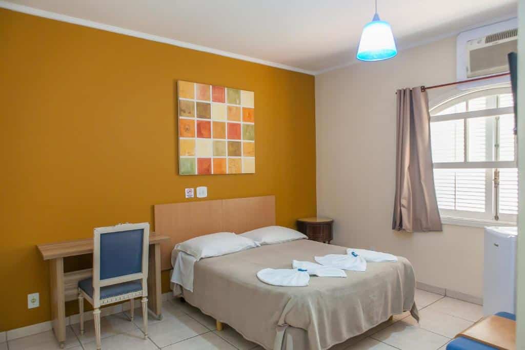 Quarto de casal na Pousada da Costa, com mesinha de trabalho, mesinha de cabeceira, frigobar, lustre e quadro numa parede amarela, para representar pousadas em Caraguatatuba.