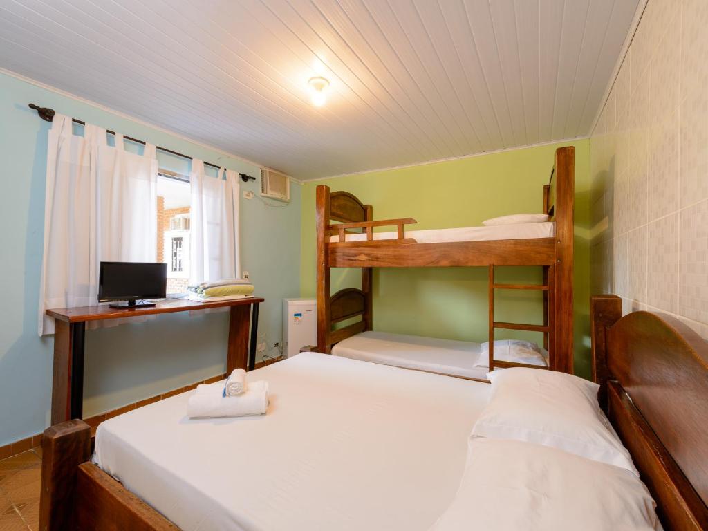 Quarto na Pousada Morada da Prainha (Prainha) com uma cama de casal, um beliche de madeira, uma escrivaninha com TV e uma janela com cortinas brancas, para representar pousadas em Caraguatatuba.