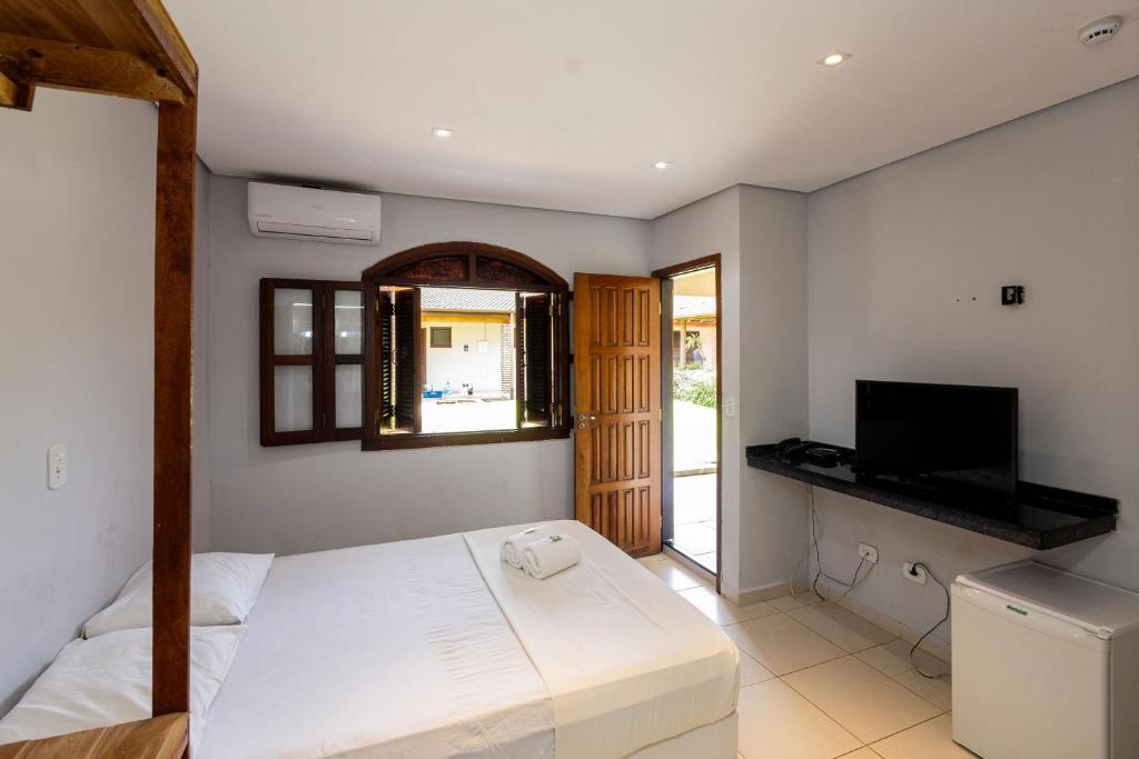 Um quarto simples na Pousada Porto do Rio (Porto Novo) com cama, TV, frigobar, janela, porta aberta e piso de azulejos brancos, para representar pousadas em Caraguatatuba.