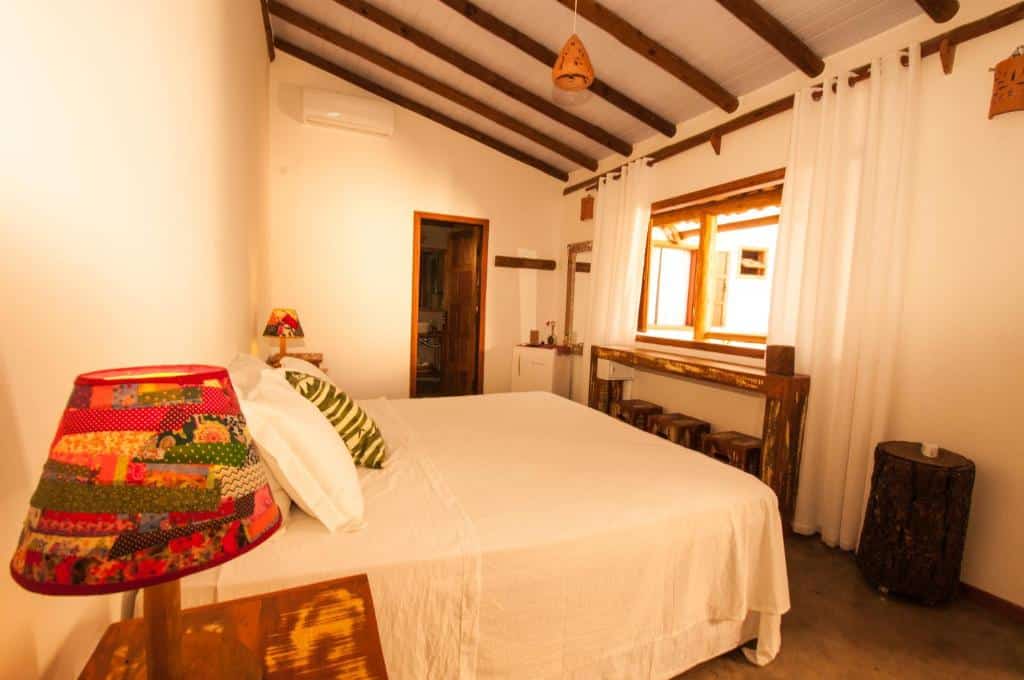 Um quarto do Hostel e Pousada Casa de Paixão Caraíva aconchegante com uma cama de casal, móveis de madeira, cortinas brancas e luminárias de patchwork em cada lado da cama.
