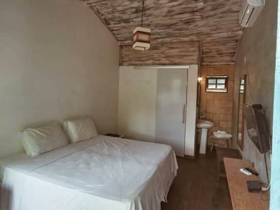 Quarto da Pousada da Praia em Caraíva com cama de casal com lençóis brancos, tv em frente e, ao lado, uma porta de correr que dá para o banheiro e há um lavado