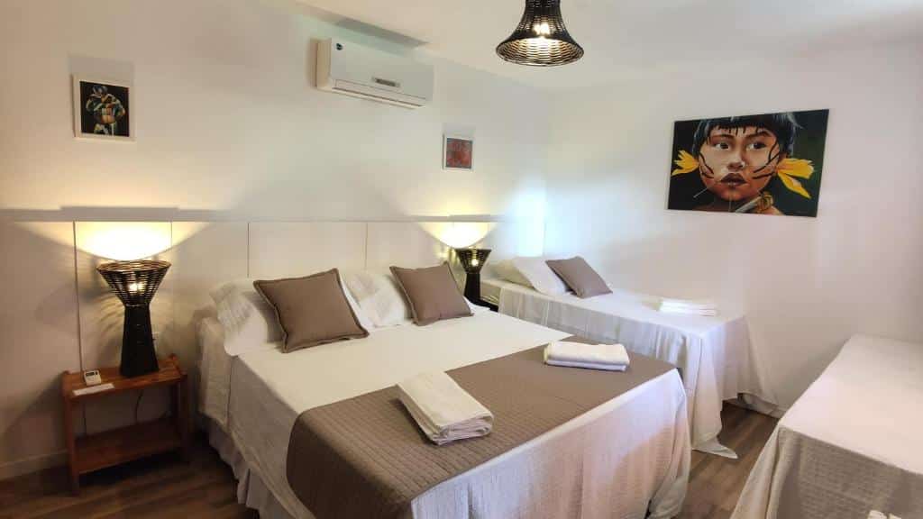 Quarto da Pousada Estrela do Mar, uma das pousadas em Caraíva com uma cama de casal e duas de solteiro, decoração neutra, arte nas paredes, luminárias e toalhas dobradas sobre as camas.