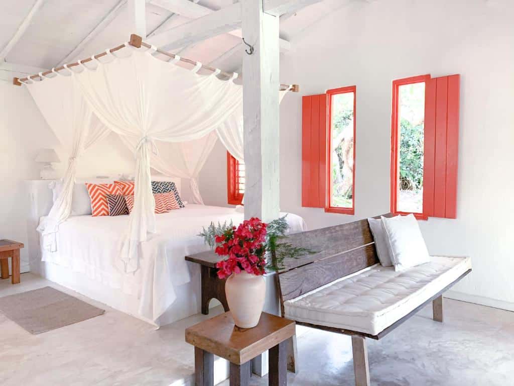 Um quarto da Pousada Lagoa bem iluminado com uma cama de dossel, persianas vermelhas, um banco de madeira com almofadas e um vaso de flores vermelhas.