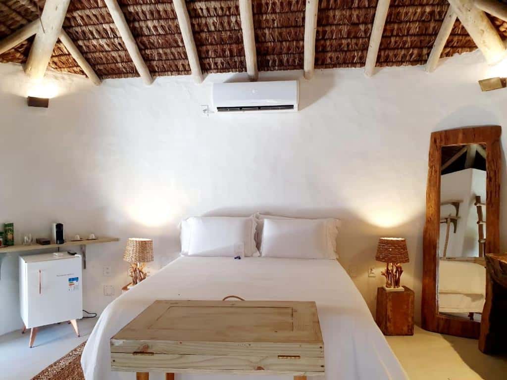 Uma cama na Villa Fulô, uma das pousadas em Caraíva, com lençóis brancos, móveis de madeira, teto de vime, um espelho de pé e um ar-condicionado na parede.