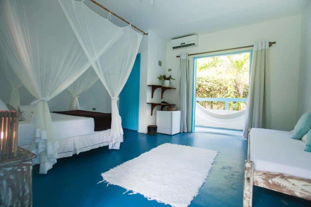 Um quarto da Pousada Caraíva bem iluminado com uma cama de dossel, tapete branco, piso azul, portas abertas para uma rede do lado de fora e uma espreguiçadeira.