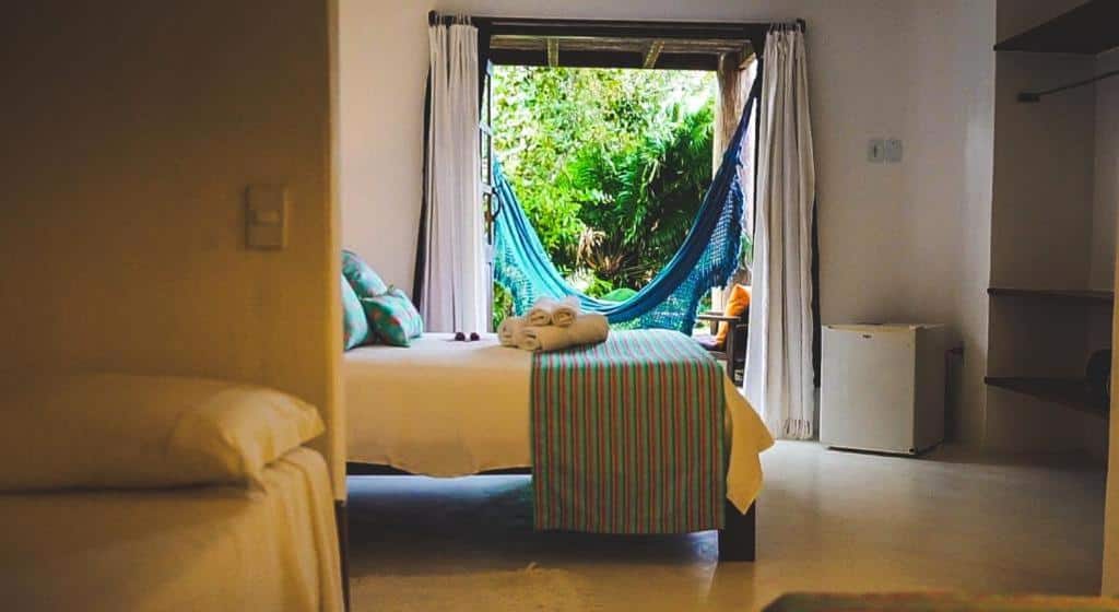 Quarto da Pousada Flor do Mar, uma das pousadas em Caraíva com uma cama com roupa de cama listrada fica de frente para uma janela com uma rede azul e vista para a vegetação; as toalhas estão dobradas sobre a cama.