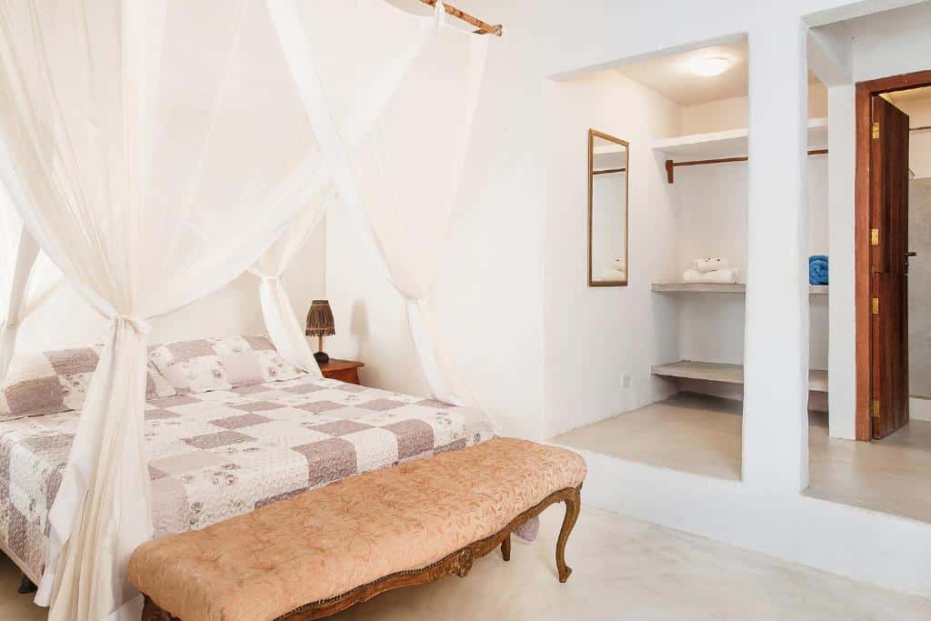 Quarto branco da Pousada Mangue Sereno, uma das pousadas em Caraíva com cama de dossel, roupa de cama floral, banco ornamentado e espaço aberto no armário com espelho e prateleiras.