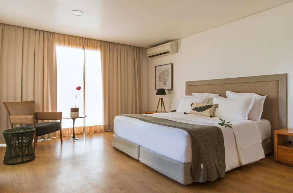 Quarto de casal na Pousada Morena, em Fernando de Noronha, com uma poltrona, um pufe e uma mesinha com orquídea, além de uma cama ampla, ar-condicionado e mesinha de cabeceira com abajur