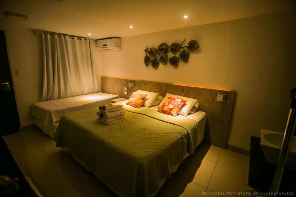 Quarto de casal com uma cama de solteiro extra na Pousada Del Mares, em Fernando de Noronha, com iluminação à meia luz, ar-condicionado no canto, cortina blecaute e enfeite na parede da cabeceira