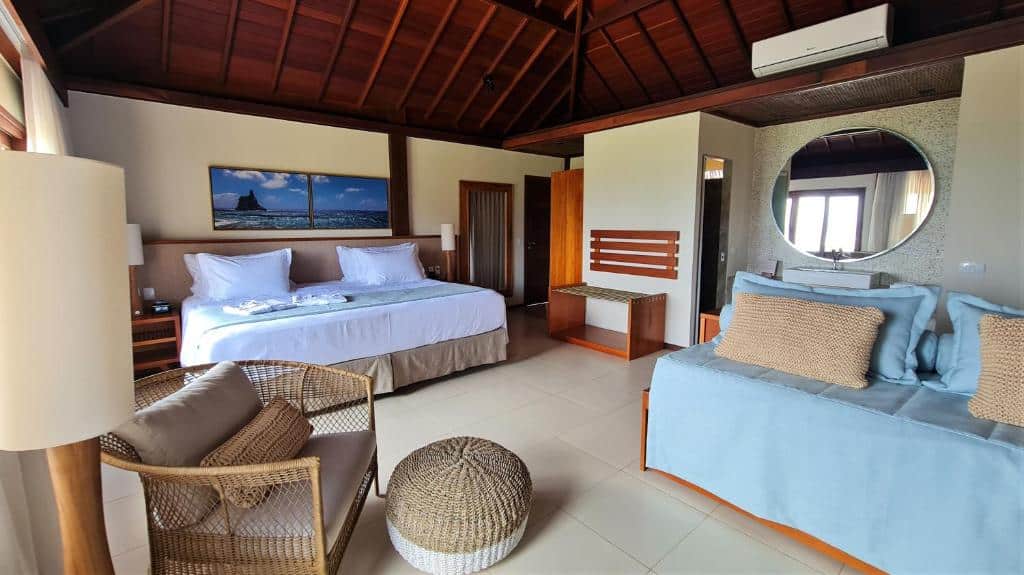 Quarto espaçoso no Nannai Solar dos Ventos, com cama ampla, poltrona, sofá, abajur de chão, quadro decorativo sobre a cabeceira, espelho redondo na parede adjacente e ar-condicionado próximo ao teto de madeira