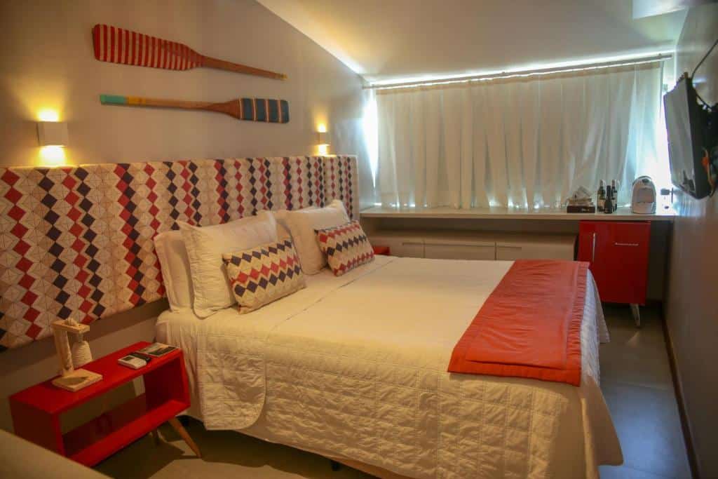 Quarto da Pousada Verdes Mares, com detalhes em vermelho e cor predominante branca, cabeceira estampada com triângulos coloridos, remos presos na parece, frigobar no fundo do quarto, cafeteira, cortina e TV