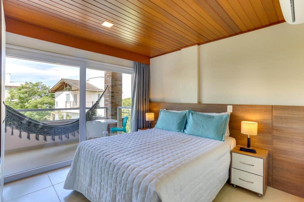 Quarto bem iluminado na Pousada Ilha Faceira com uma cama de casal, duas mesinhas de cabeceira, luminárias e uma rede na varanda do lado de fora da porta de vidro deslizante, para representar pousadas em Florianópolis.