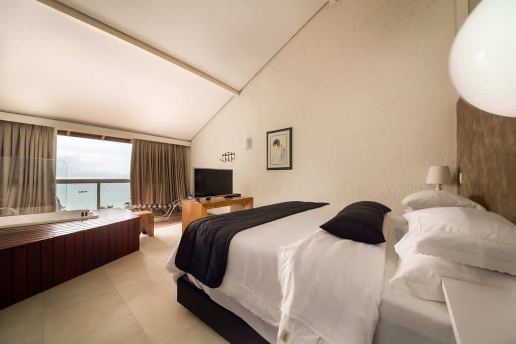 Quarto na Pousada dos Sonhos com cama de casal, TV, jacuzzi perto da janela e vista para o oceano através de grandes portas de vidro, para representar pousadas em Florianópolis.