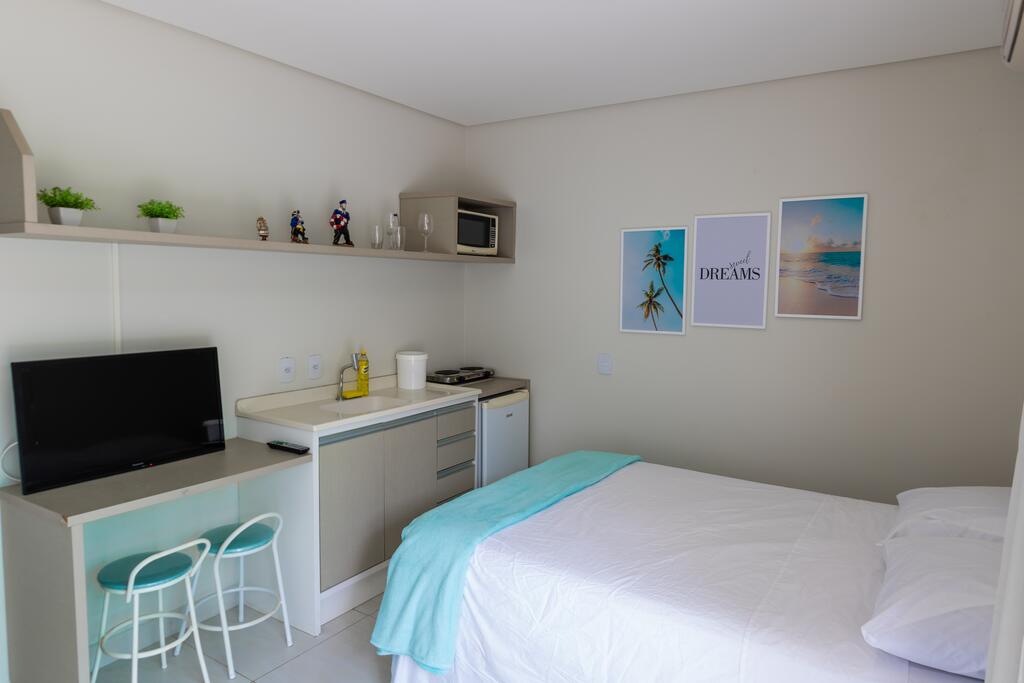 Um pequeno estúdio na Pousada Dona Aurea com uma cama, cozinha compacta, TV, duas banquetas e quadros emoldurados em uma parede bege, para representar pousadas em Florianópolis.
