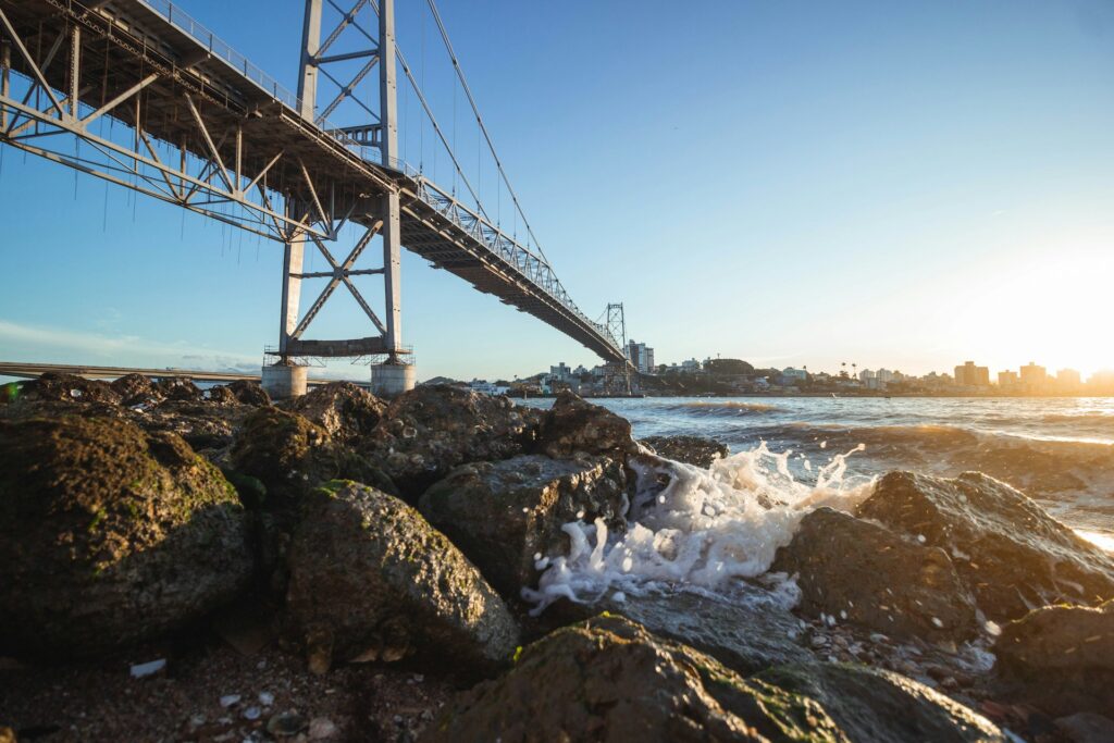 Vista de ângulo baixo de uma grande ponte de metal sobre a água, com ondas batendo em rochas e prédios da cidade de Florianópolis ao fundo.