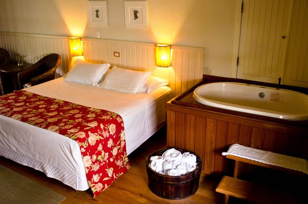 Um quarto na Pousada Vila Tamarindo Eco Lodge com uma cama de casal, um tapete de cama floral, luzes de cabeceira, uma banheira e uma cesta de toalhas no chão, para rperesentar Pousadas em Florianópolis.