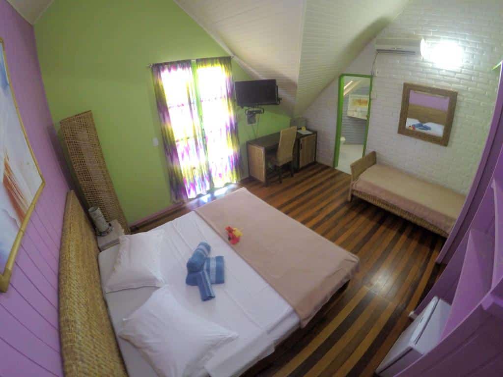 Um quarto arrumado na Pousada Rio Vermelho com uma cama de casal, uma pequena escrivaninha, um sofá-cama e piso de madeira listrado, visto de cima, para representar pousadas em Florianópolis.