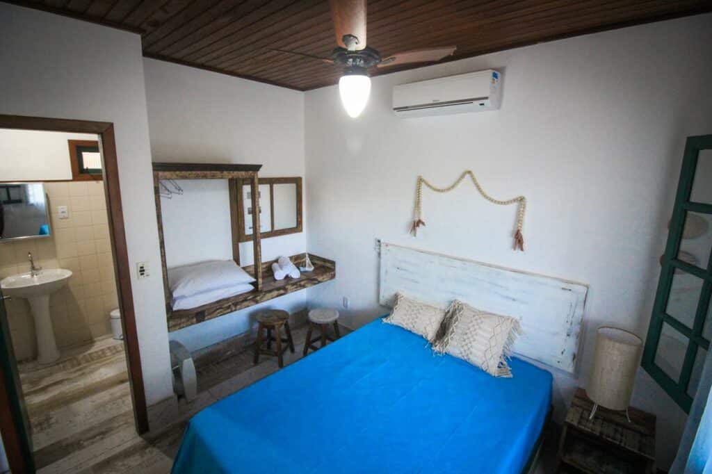 Quarto pequeno na Vila Campeche com colcha azul, móveis rústicos de madeira e uma entrada aberta para o banheiro à esquerda, para representar pousadas em Florianópolis.