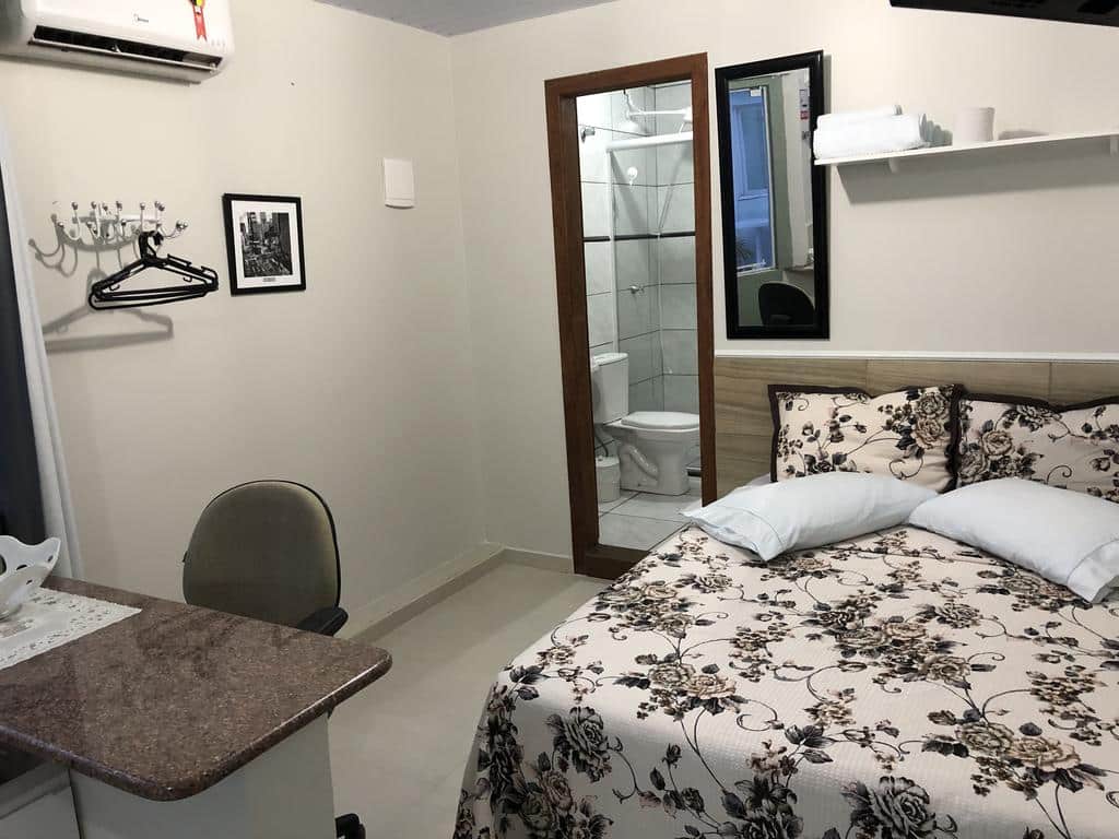 Quarto pequeno na Pousada Marques Floripa com colcha floral, escrivaninha e cadeira, ganchos de parede e vista para um banheiro de azulejos com vaso sanitário, para representar pousadas em Florianópolis.