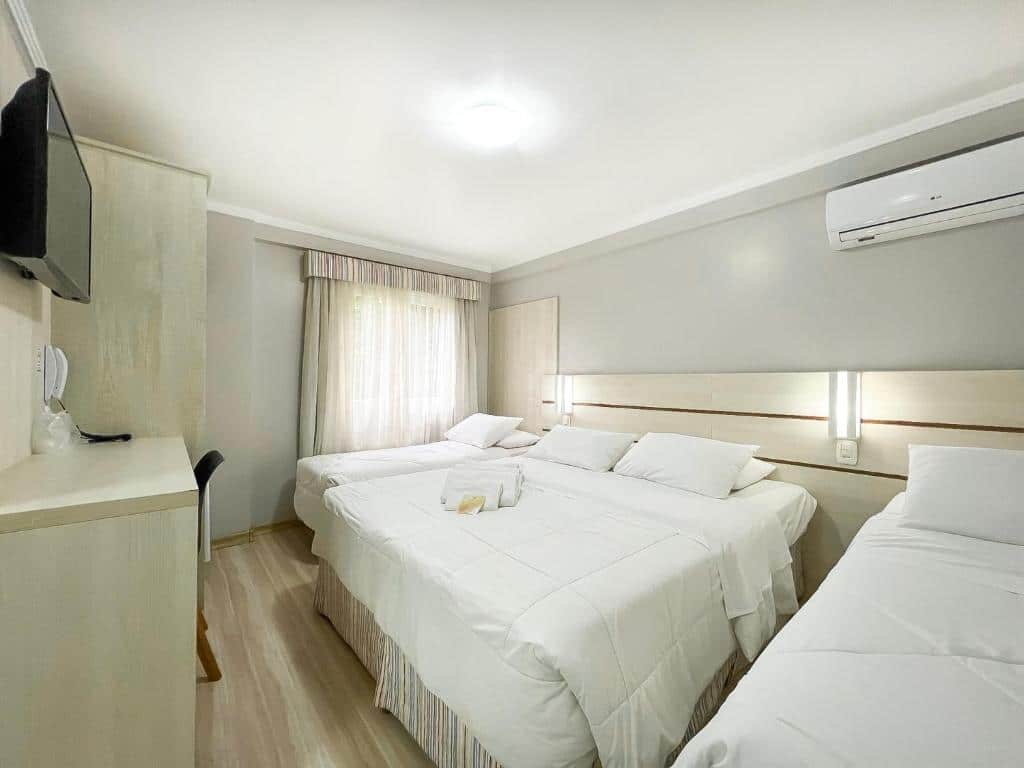 Quarto da Pousada Viena com três camas dispostas lado a lado, uma de casal ao centro e duas de solteiro. No quarto também há uma televisão presa à parede no lado esquerdo e um ar-condicionado sob a cama, no lado direito, para representar pousadas em Gramado.