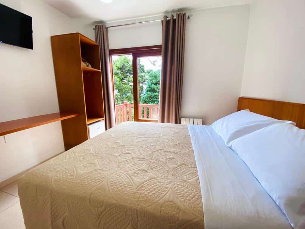 Quarto da Pousada do Lago com uma cama de casal no lado direito e uma porta para a sacada no canto esquerdo. A visão lá de fora é de várias araucárias, na propriedade da Pousada do Lago Gramado.