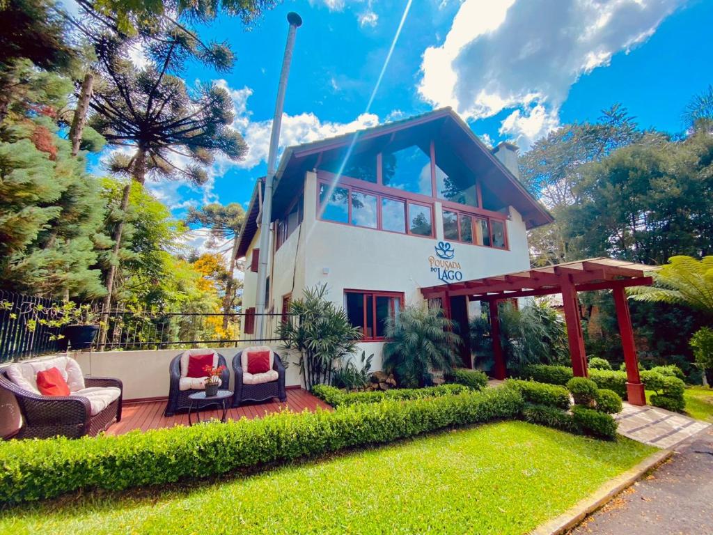 Vista frontal da Pousada do Lago com um belo jardim cuidado na entrada, uma área de descanso com três poltronas estofadas ao ar livre e um céu azul com poucas nuvens ao fundo. Ao redor da pousada há diversas árvores araucárias.