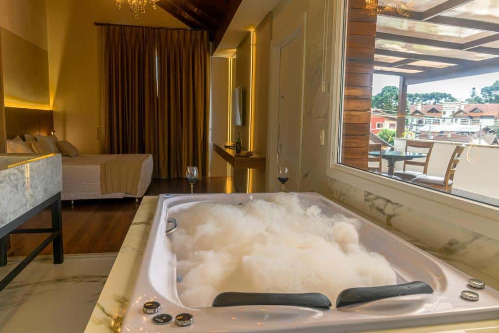 Suíte da Pousada Kaster com uma bela banheira de hidromassagem, cheia de espuma e com duas taças com vinho postas em frente a banheira. Mais ao fundo é possível ver uma cama de casal à esquerda do quarto, e pela janela no canto direito, é possível ver a sacada mobiliada da suíte com uma mesa redonda e algumas cadeiras ao redor, para representar pousadas em Gramado.
