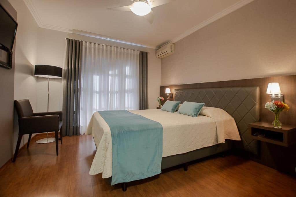 Quarto da Pousada Vovó Carolina com uma cama de casal decorada com lençol e travesseiros azuis claros. No canto esquerdo do quarto há uma cadeira de courino e uma televisão presa à parede, para representar pousadas em Gramado.