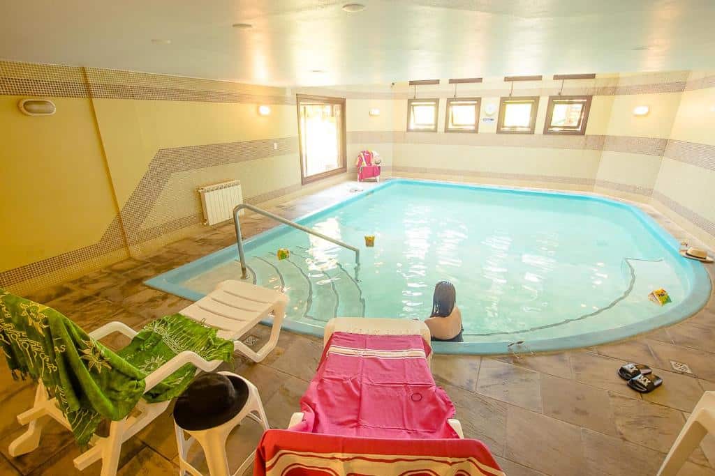 Piscina aquecida da Pousada Vovó Carolina com uma mulher sentada dentro dela, de costas para a câmera. Ao redor da piscina há duas espreguiçadeiras de plástico, brancas, com toalhas coloridas esticadas sobre elas.