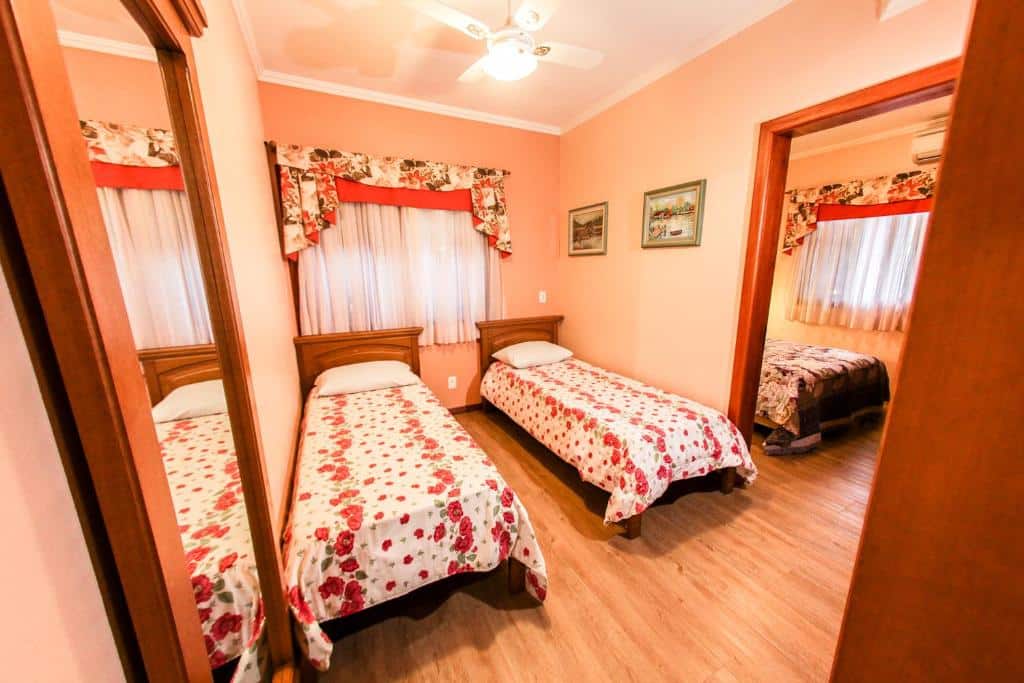 Quarto da Pousada das Papoulas em Gramado com duas camas de solteiro decoradas com lençóis floridos em tons vermelhos, e uma porta à direita com acesso ao quarto ao lado mostrando uma parte de uma cama de casal.